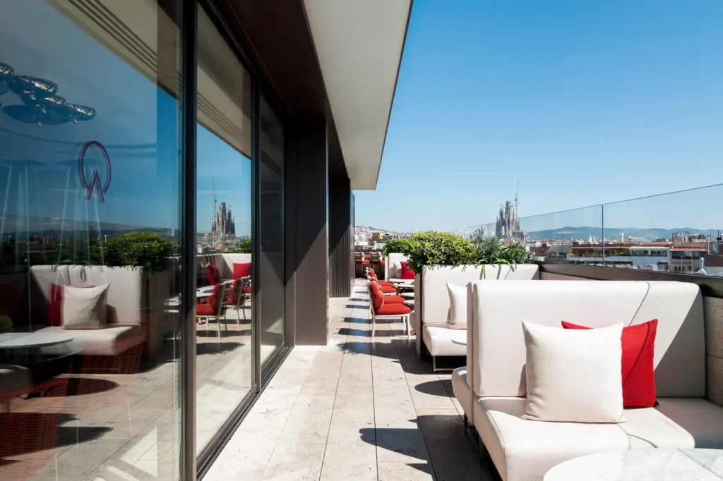Moderna terraza en la azotea con asientos blancos acolchados, cojines rojos y vistas a la ciudad bajo un cielo azul despejado; una pared de cristal refleja el elegante espacio y la piscina cercana.